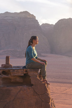 the girl-traveler sits on a stone ledge against the backdrop of the mountains in the Wadi Wadi desert at sunset