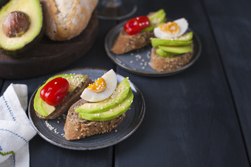 Tasty sandwich with avocado, tomato and poached egg on wooden chopping board, close up, selective focus. Healthy delicious breakfast or lunch. On a dark background, top view