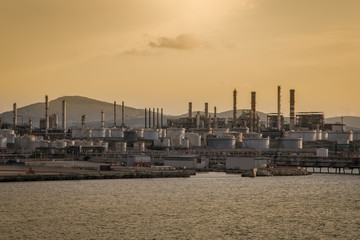 Obraz premium Skyline industrial area Cagliari, Sardinia Italy. View from the sea of an oil refinery plant from industry zone with cloudy sky at sunset. Oil storage tank in port area.