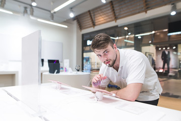 Handsome man stands next to a showcase with tablet stylus in his hands and looks at the camera. Buyer looks at the tablets in the electronics store.