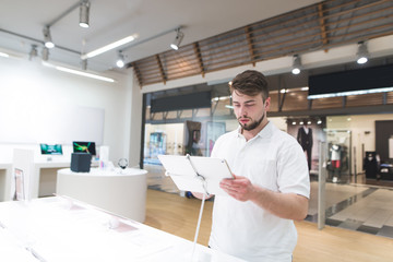 Handsome man stands with a tablet in his hands in a modern tech store. Portrait of the buyer of the...