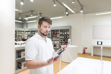 Portrait of a handsome man standing with two tablets in the hands of a tech store background and looking into the camera. Buyer chooses a tablet in the electronics store.