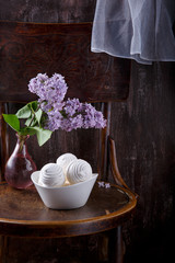 Bowl of white zephyr marshmallows and bouquet of lilac flowers on old vintage chair.  Still life on dark background.