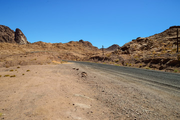 mountain desert landscape, mountains of red sandstone, a plain covered with rare desert vegetation, a stretch of road with telegraph poles against the background of a cloudless blue sky, Southern Sina