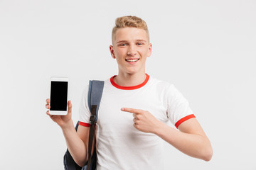 Image of happy male student having clean healthy skin wearing backpack pointing finger at black copyspace screen of smartphone, isolated over white background