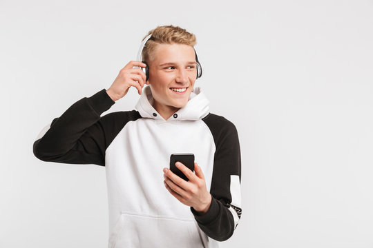 Portrait of caucasian youngster 16-18 years old wearing hoodie smiling and listening to music on mobile phone via wireless earphones, isolated over white background