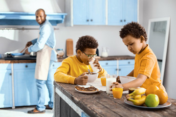 Morning routine. Charming little boys sitting at the table and having breakfast while their father cooking in the background