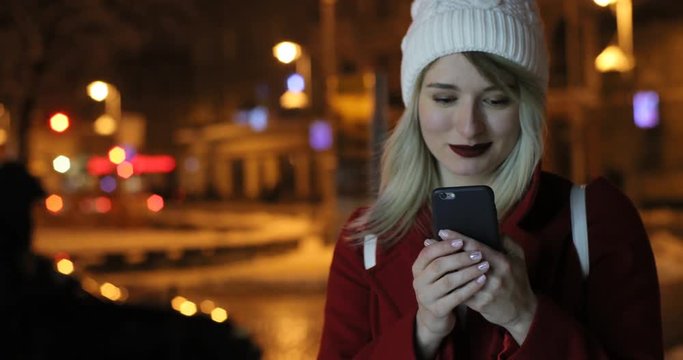 Outdoor portrait of beautiful young woman using her mobile phone at night street. Winter, festive Christmas fair on background.