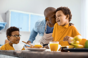Overwhelmed with feelings. Affectionate young father kissing his little son on the cheek while he having breakfast in the kitchen