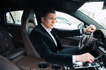 Real businessman! Side view portrait of the handsome brutal serious young businessman sitting in the car and driving to his huge modern office.