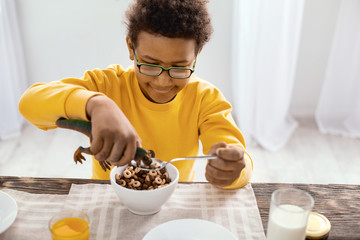 Hungry toy. Upbeat pre-teen boy sitting at the table and feeding cereals to toy dinosaur while...
