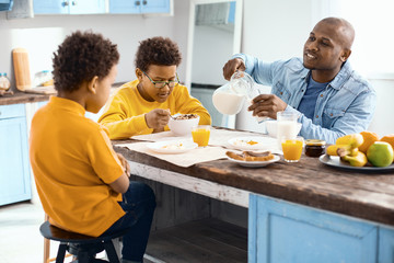 Fototapeta na wymiar Discussing plans. Upbeat young man pouring a glass of milk and chatting with his sons while they all having breakfast in the kitchen