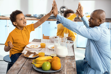 Best team. Cheerful single-parent family sitting at the kitchen table and giving each other a high five while having breakfast