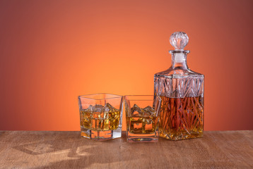 Old fashion glass and carafe with the alcohol, whisky drink on warm, dark orange background and wood
