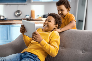 Brotherly care. Loving little boy giving his elder brother massage while the elder boy sitting on the sofa and playing a game on tablet