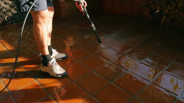 Male in shorts cleans outdoor terrace tiles with high pressure washer. Water sprinkling and shining in evening sunset. Spring cleaning arround the house.