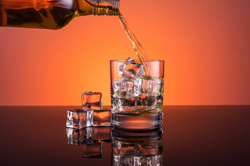 Whisky alcohol pouring into glass with ice drink on warm dark orange background