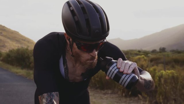 Tracking video shot of professional cyclist riding bike fast and drinking water from and pouring water on his head. Racer getting hydrated while riding a bicycle on outdoors.