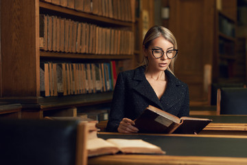 Beautiful blonde woman wearing elegant black tweed jacket and glasses sitting at desk beside bookshelf and reading book. Young gorgeous female student studying at library. Smart is new sexy concept.