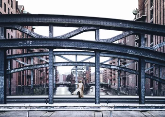 Rückansicht einer Frau mit langem Mantel, die auf einer windigen Brücke in der alten Speicherstadt in Hamburg steht © Christian Horz