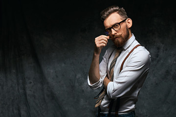 Young stylish hipster with cool hairstyle and beard dressed in white shirt and suspenders is...