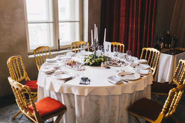 Round table with many chairs around served for many people in retro style with white tablecloth, luxurious cultery and candlestick in middle at antique restaurant. Table set for business people
