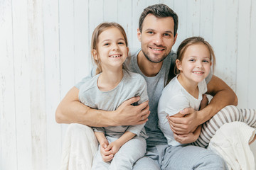 Portrait of happy young father embraces his two daughters, loves them very much, pose together at...