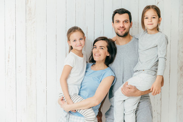 Portrait of happy parents carry their beautiful daughters on hands. Lovely family of four pose together against white background have positive expressions. Affectionate mother and father with children