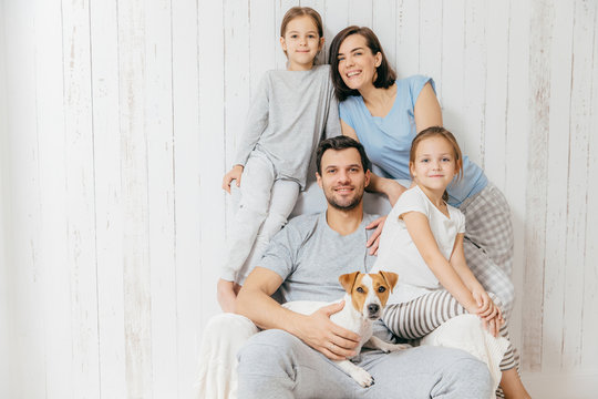 Horizontal shot of friendly family pose together against white background: two little sisters, father, mother and their pet. Happy parents and their female children. Family of four. Parenthood