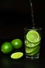 Cold detox water with ice and fresh ripe slice green limes on black background.