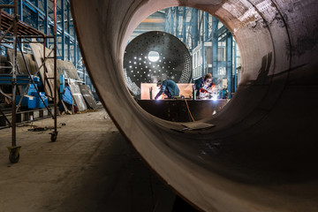 Two workers welding in the interior of a factory manufacturing metallic cylinders for industrial...