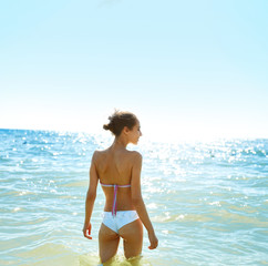 Fototapeta na wymiar beautiful sexual young woman in white bikini stands on the beach against the sea and sky at sunny day. happy woman enjoys summer vacation and goes to swim in the sea. Back view