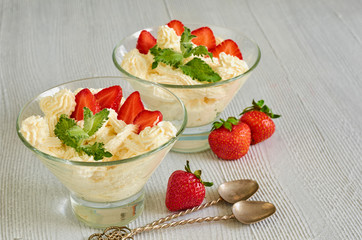 Cold summer dessert with sliced strawberries, cheese cream in the glass bowls on the gray kitchen table. Parfait decorated with fresh mint leaves and two vintage spoons. Side view