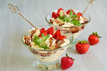 Parfait with sliced strawberries and chocolate splash in the glass bowls on the gray kitchen table. Cold summer dessert decorated with fresh mint leaves and two vintage spoons. Close up view