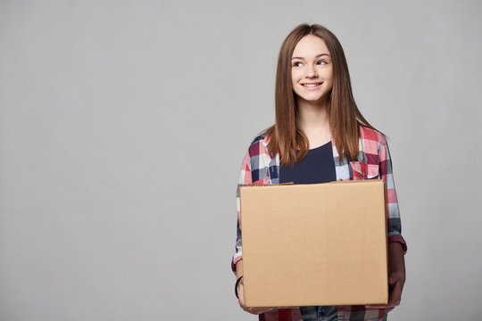Delivery, relocation and unpacking. Smiling young woman holding cardboard box looking to side at blank copy space, over grey background