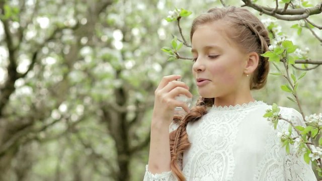 Girl with braid wearing in a white dress sniffing perfumes in bottle and blossoming branch in the pear garden in slow motion