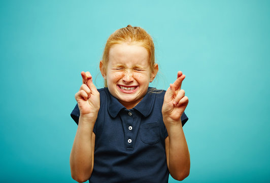 Children girl tightly closed his eyes and put fingers crossed, make a wish, believe in the dream, expresses heartfelt emotions, has funny facial features, beautiful red hair with freckles.