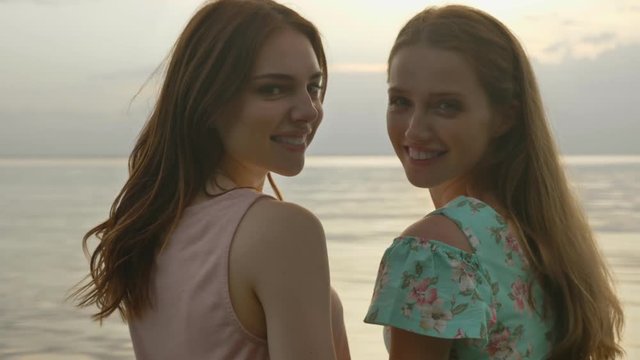 Two young Caucasian girls in dresses walking along shallow water at sunset, turn around and look at the camera