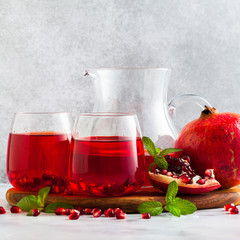 freshly prepared pomegranate juice in a decanter and two glasses on a marble table and a gray stone background with mint leaves and fruit seeds