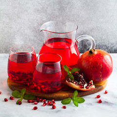 freshly prepared pomegranate juice in a decanter and two glasses on a marble table and a gray stone background with mint leaves and fruit seeds