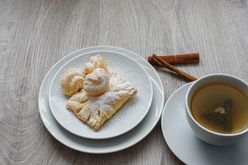 Puff pastry with sweet filling with powdered sugar on a white plate cup of tea and vanilla sticks on a wooden background