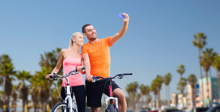 fitness, sport, technology and healthy lifestyle concept - happy couple riding bicycles and taking selfie by smartphone over venice beach background in california