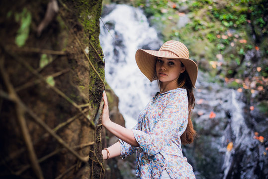 Beautiful young woman standing at the lake near waterfall in white dress and straw hat