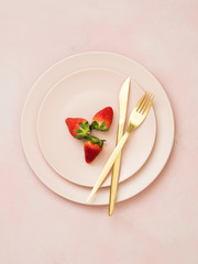 Top view of ceramic plates with strawberries and golden cutlery on pink pastel background