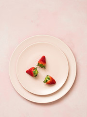 Top view of ceramic plates with three strawberries on pink pastel background