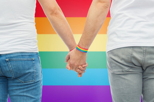 lgbt, same-sex relationships and homosexual concept - close up of male couple wearing gay pride awareness wristbands holding hands over rainbow colors background
