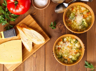 Two bowl of minestrone soup with cheese on cutting board and vegetables on rustic wooden background, top view.