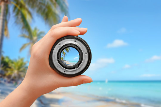 Female hand holding lens on the beach, palm trees and sea in the background. Travel and photography concept