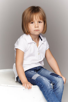 Portrait of an upset little girl in denim overalls, sitting in a studio on gray background.