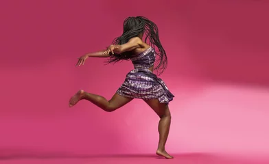 Tuinposter Beautiful African Black girl wearing traditional colorful African outfit does a dramatic dance move against a colorful pink background © Paul